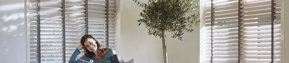Vrouw die ontspannen zit onder openstaande houten jaloezieën in een woonkamer