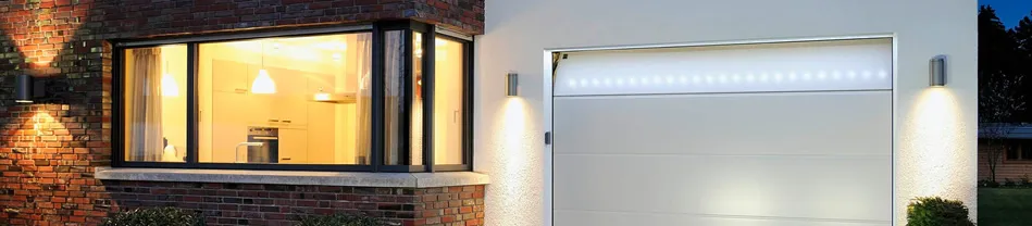 Wit garagepoortdeur naast een verlichte keukenruimte met grote ramen en buitenverlichting
